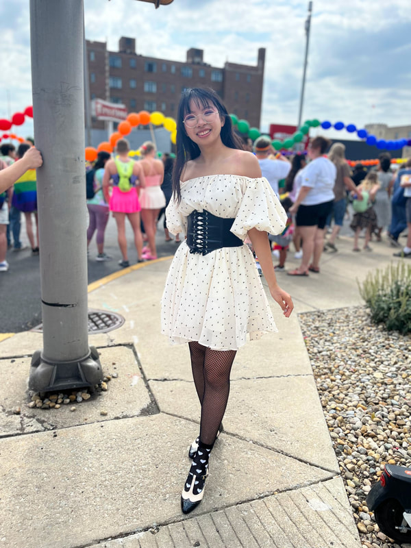 me wearing a poofy ivory mini dress with a corset belt, posing in front of a pride parade