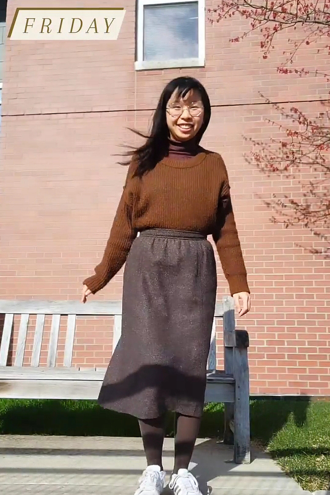 grainy screenshot of me wearing a brown turtleneck, brown sweater, brown A-line skirt, and brown tights standing in front of a brick building